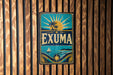 A vintage-style metal sign with the word "EXUMA" prominently displayed, featuring a sun, palm trees, and a sailboat.
