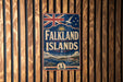 A vintage-style metal sign with the text "Falkland Islands" and a Union Jack flag, mounted on a wooden wall.