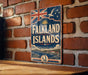 A vintage-style sign for Falkland Islands, featuring a Union Jack flag and a seagull, is displayed on a brick wall.