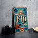 A vintage-style metal sign with the word "Fiji" prominently displayed, featuring a tropical landscape with palm trees, a sun, and a boat.