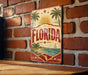 A vintage-style metal sign with the word "FLORIDA" prominently displayed, featuring palm trees, a sailboat, and a sun-filled sky.