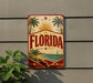 A vintage-style sign with the word "FLORIDA" prominently displayed, featuring palm trees, a sun, and a sailboat, all set against a beach and ocean backdrop.