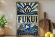 a vintage-style sign with a blue and white color scheme, displaying the word "FUKUI" prominently. The sign also includes a depiction of a Japanese castle and a seascape, suggesting a connection to Japanese culture and nature. The sign is placed on a wooden surface, with a potted plant and a wooden cutting board visible in the background.