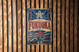 a vintage-style sign with the word "FUKUOKA" prominently displayed, along with a graphic of a temple and a sun. The sign is mounted on a wooden wall with vertical slats.