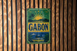 A vintage metal sign with the word "GABON" displayed prominently in the center, surrounded by a tropical landscape with palm trees and a sun.