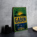 A vintage metal sign with the word "GABON" prominently displayed, featuring a tropical landscape with palm trees and a sun.