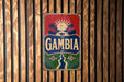 A vintage-style sign with the word "GAMBIA" prominently displayed, featuring a tropical landscape with palm trees and a sun.