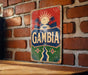 a vintage-style sign with the word "GAMBIA" prominently displayed, mounted on a brick wall.