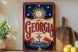 A vintage-style metal sign with the word "Georgia" prominently displayed, featuring a sun and stars design, is mounted on a wall next to a wooden cutting board and some kitchen utensils.
