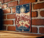 A vintage-style metal sign with the word "GEORGIA" prominently displayed, featuring a sun and stars design, is mounted on a brick wall.