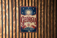 A vintage-style metal sign with the word "GEORGIA" prominently displayed, featuring a sun and stars design, is mounted on a wooden wall.