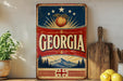 A vintage-style sign with the word "GEORGIA" prominently displayed, featuring a peach and stars, along with a mountain landscape and a Georgian flag. The sign is placed on a shelf next to a wooden cutting board and some lemons.