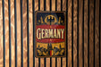 A vintage-style metal sign with the word "GERMANY" prominently displayed, featuring an eagle and a cityscape design.