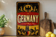 a vintage-style sign with the word "GERMANY" prominently displayed, surrounded by various German-themed elements such as a crest, buildings, and a sun. The sign is placed on a shelf next to a wooden cutting board and a potted plant, creating a rustic and cozy atmosphere.