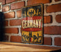 A vintage-style metal sign with the word "GERMANY" prominently displayed, featuring an illustration of a city skyline and a sun.