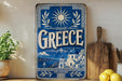 a vintage-style metal sign with a blue background and white text, depicting a scenic view of Greece. The sign is placed on a wooden surface, surrounded by various kitchen utensils and a potted plant.