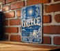 A vintage metal sign with the word "Greece" prominently displayed, featuring a sun and laurel wreath design, mounted on a brick wall.