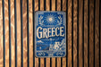 A vintage-style metal sign with the word "Greece" prominently displayed, featuring a sun and laurel wreath design.