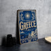 A vintage-style metal sign with the word "Greece" prominently displayed, featuring a sun and olive tree design, is leaning against a concrete wall.
