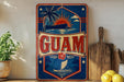 a vintage-style sign with the word "GUAM" prominently displayed, surrounded by various tropical elements such as palm trees, a sunset, and a boat.