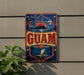 a vintage-style sign with the word "GUAM" prominently displayed, along with a palm tree and a sailboat, all set against a blue background with a red border.