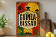 a colorful sign with the text "GUINEA-BISSAU" prominently displayed, surrounded by various tropical and cultural elements such as palm trees, a star, and a boat.