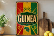 a vintage-style metal sign with the word "GUINEA" prominently displayed, surrounded by a colorful design with a sun and stars. The sign is placed on a shelf next to a wooden cutting board and a potted plant.