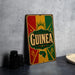 a vintage-style sign with the word "Guinea" prominently displayed, along with a map of the country. The sign is placed on a gray surface, and there are some black objects, such as a stack of cups, to the left of the sign.