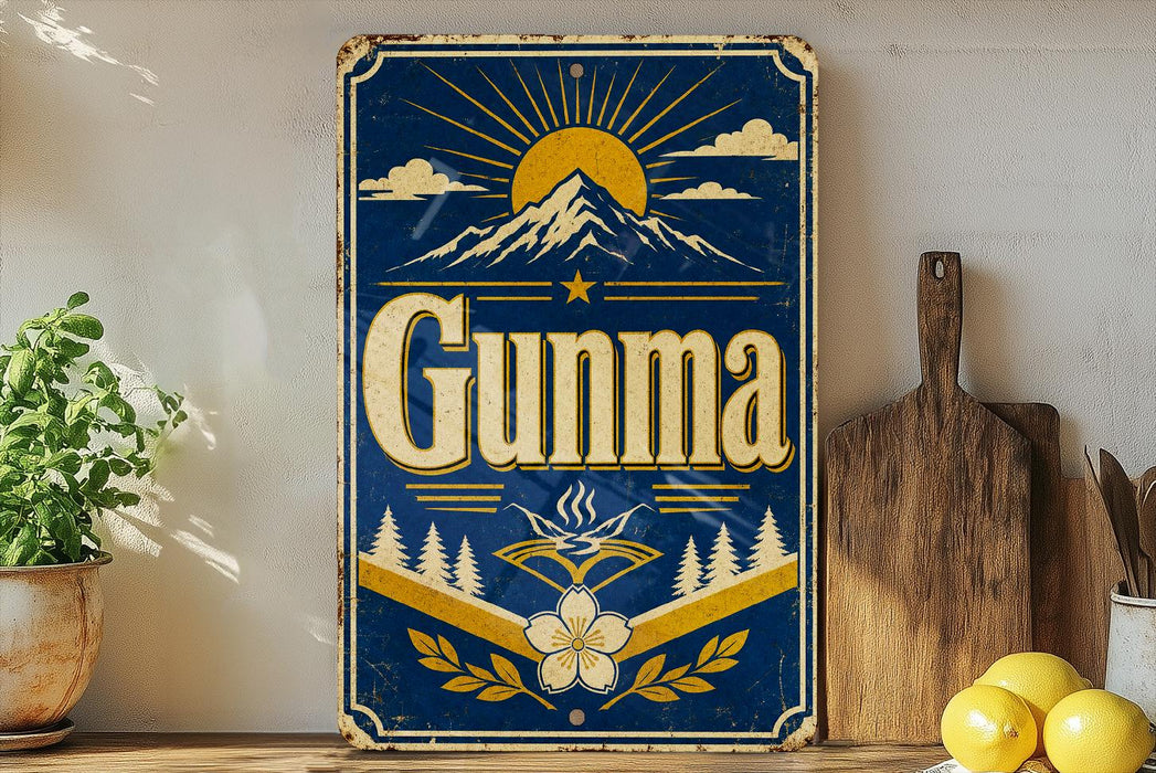 a vintage-style sign with the word "GUNMA" prominently displayed, surrounded by a mountain landscape and sun-like rays. The sign is placed on a wooden surface, with a potted plant and a wooden cutting board visible in the background.