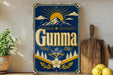 a vintage-style sign with the word "GUNMA" prominently displayed, surrounded by a mountain landscape and sun-like rays. The sign is placed on a wooden surface, with a potted plant and a wooden cutting board visible in the background.