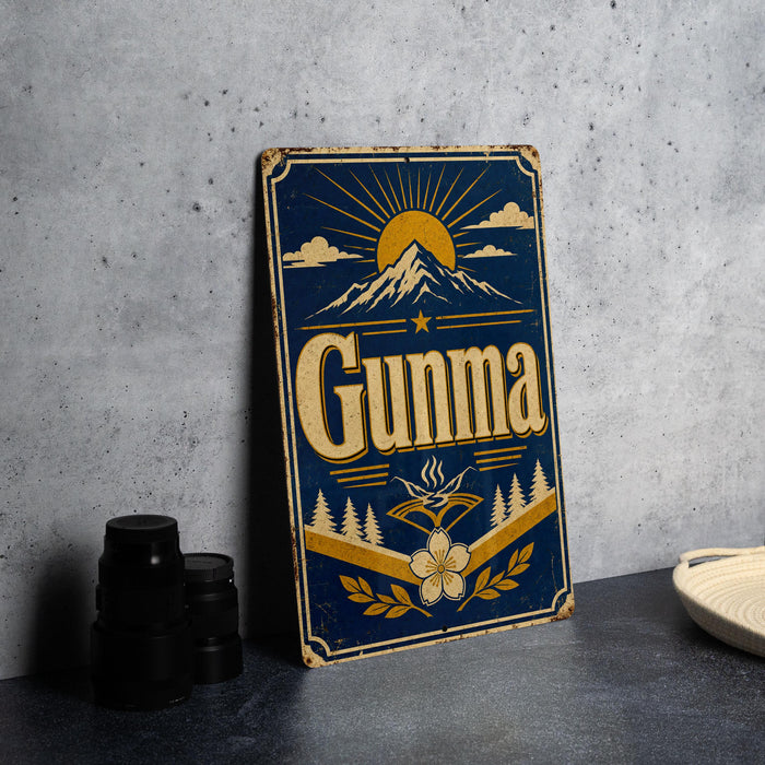 A vintage metal sign with the word "GUNMA" prominently displayed, featuring a mountain landscape and sun design.