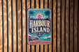 A vintage-style sign for Harbour Island, featuring a palm tree, sun, and other nautical elements, is displayed on a wooden wall.