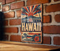 A vintage-style metal sign with the word "HAWAII" prominently displayed, featuring a sun and palm trees, along with other tropical elements.
