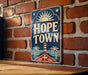 A vintage-style metal sign with the text "HOPE TOWN" displayed prominently on a brick wall.