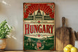 A vintage-style metal sign with the word "Hungary" prominently displayed, featuring a castle and bridge in the background. The sign is placed on a shelf next to a wooden cutting board and some potted plants.