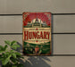 A vintage-style metal sign with the word "Hungary" prominently displayed, featuring a castle and other architectural elements. The sign is mounted on a wall, and there is a plant visible to the left of the sign.