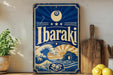 A vintage-style sign with the word "Ibaraki" prominently displayed, featuring a wave and a sun design. The sign is placed on a wooden surface, with a potted plant and a wooden cutting board nearby.