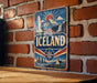 A vintage-style sign for Iceland is displayed on a brick wall, featuring a mountain landscape and a sunburst design.