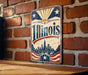 A vintage-style sign displaying the word "Illinois" in large letters, with a cityscape and sun motif in the background.