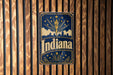 A vintage-style metal sign with the word "Indiana" prominently displayed, featuring a flame and stars against a dark blue background.