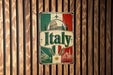 A vintage-style metal sign with the word "Italia" prominently displayed, featuring a combination of green, red, and white colors, along with images of iconic landmarks and symbols associated with Italy.