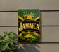A vintage-style metal sign with the word "Jamaica" prominently displayed, featuring a tropical island landscape with palm trees and a sunburst design.
