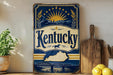 A vintage-style sign for Kentucky featuring a sun and stars design, with the word "Kentucky" prominently displayed. The sign is placed on a wooden surface, with a potted plant, a wooden cutting board, and some lemons nearby.