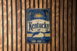 A vintage-style metal sign with the word "Kentucky" prominently displayed, featuring a sun and a horse, is mounted on a wooden wall.