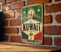 A vintage-style sign with the word "KUWAIT" prominently displayed, featuring a city skyline and a palm tree. The sign is mounted on a brick wall, and there is a wooden table below it.