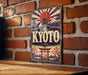A vintage-style sign with the word "KYOTO" prominently displayed, featuring a red sun, a temple, and other traditional Japanese elements. The sign is mounted on a brick wall, and there is a wooden table in the foreground.