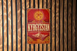 A vintage-style sign with the word "KYRGYZSTAN" displayed prominently in the center, surrounded by a sun-like symbol and mountainous landscape.