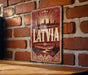 A vintage-style sign with the word "LATVIA" prominently displayed, featuring a sunburst design and silhouetted cityscape.