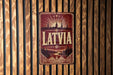 A vintage metal sign with the word "LATVIA" prominently displayed, featuring a scenic landscape with a river and mountains in the background.