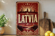 A vintage-style metal sign with the word "LATVIA" prominently displayed, featuring a sunburst design and silhouettes of buildings and trees. The sign is placed on a wooden surface, with a potted plant, a wooden cutting board, and some lemons nearby.